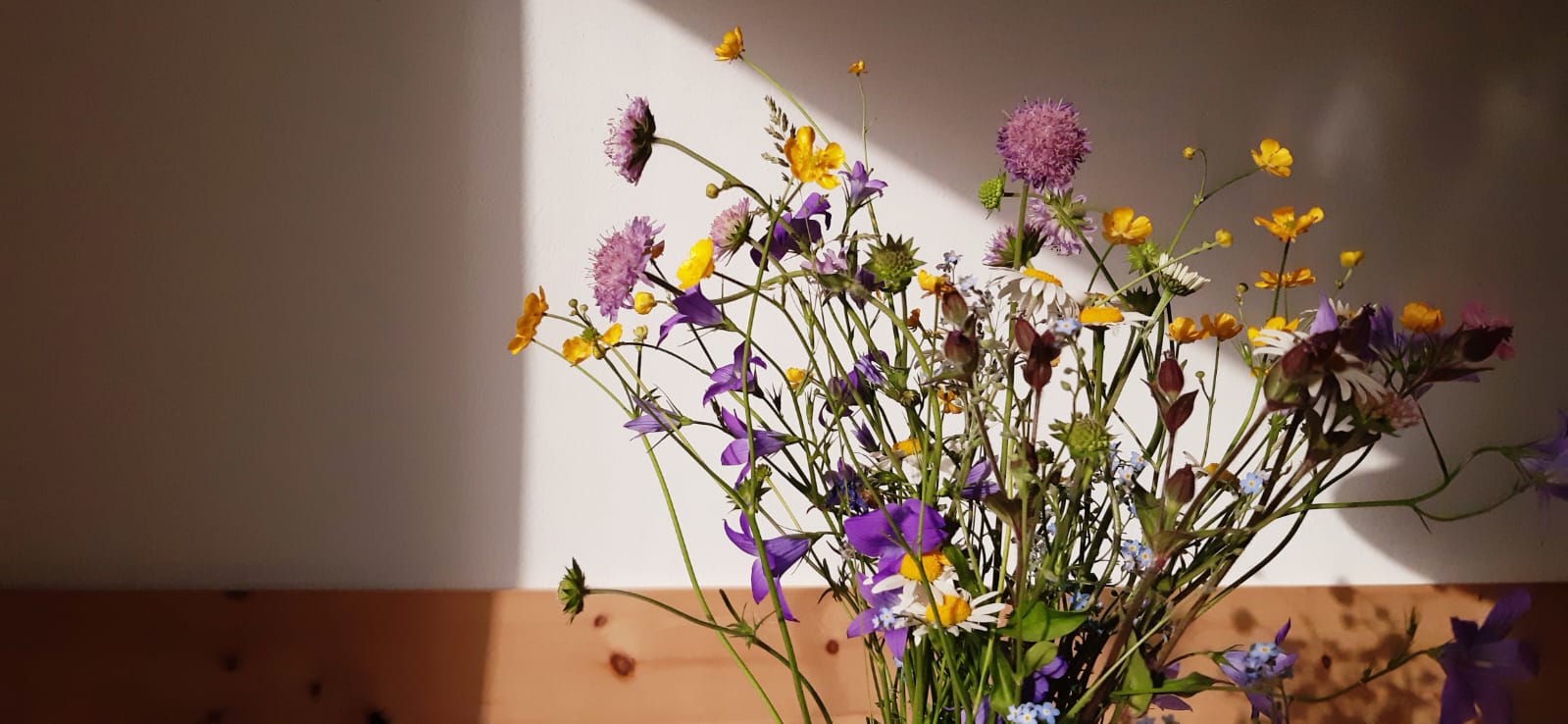 Ein bunter Blumenstrauß vor einer teils verputzten, teils mit Holz verkleideten Wand, auf die ein dreieckiger Lichtfleck fällt